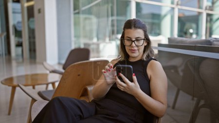 Téléchargez les photos : Femme hispanique rayonnante, tout sourire, messages animés sur son smartphone tout en se prélassant à la mode dans un hall d'hôtel moderne - en image libre de droit