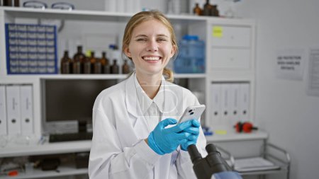 Téléchargez les photos : Souriante jeune femme caucasienne en blouse blanche à l'aide d'un smartphone dans un cadre de laboratoire. - en image libre de droit