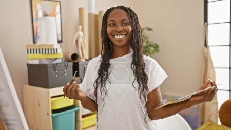 Foto de Mujer afroamericana artista sonriendo en un estudio creativo interior sosteniendo pinceles - Imagen libre de derechos