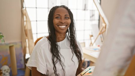 Foto de Retrato de una joven sonriente con el pelo rizado, vestida artísticamente, en un entorno de estudio de arte iluminado por el sol. - Imagen libre de derechos