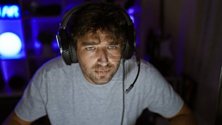 A handsome man with green eyes and a beard wearing headphones in a dark gaming room at night, looking focused and engaged.