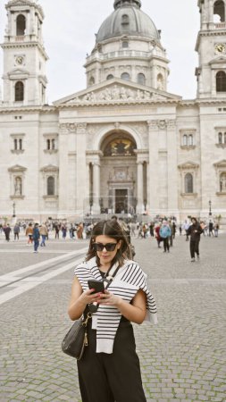 Foto de Una mujer latina con estilo utiliza un teléfono inteligente frente a la basílica de st. stephen de budapest - Imagen libre de derechos