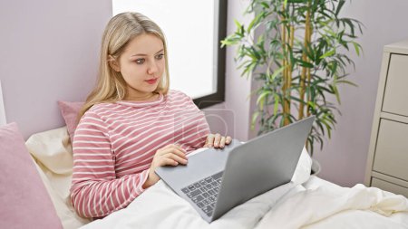A young blonde caucasian woman working on a laptop in a cozy bedroom with plants in the background.
