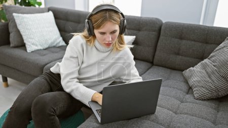 Foto de Una mujer rubia enfocada que usa auriculares usa una computadora portátil en un sofá en una sala de estar moderna. - Imagen libre de derechos