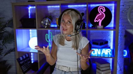 Photo for In the heart of a virtual night, a young, attractive blonde woman streamer, headset-on, steadily speaking during a live game stream in her dark, futuristic gaming room - Royalty Free Image