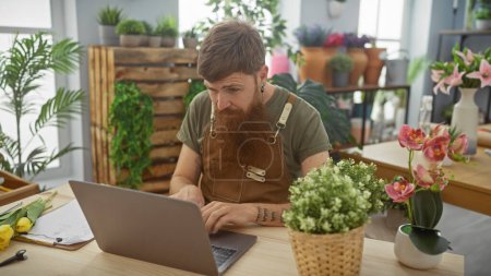 Redhead bearded man working on laptop in a flower shop with plants in background.
