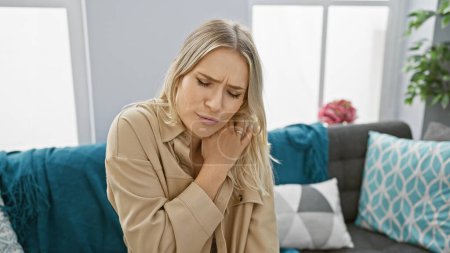 Worried and unhappy, young blonde woman suffers severe cervical pain sitting alone on sofa at home
