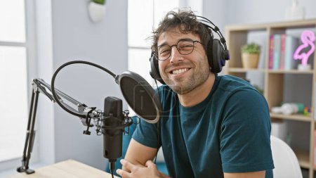 Lächelnder junger hispanischer Mann mit Kopfhörer, der ein Mikrofon in einem hellen Innenstudio bedient.