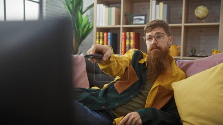 Bearded man lounging on couch with remote in a cozy home interior.