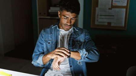 Photo for Hispanic man in casual attire looking at smartwatch in a modern office setting - Royalty Free Image