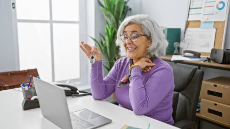 Photo for A cheerful woman with grey hair presenting keys in a bright office setting, embodying professional success and satisfaction. - Royalty Free Image