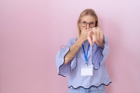 Junge kaukasische Geschäftsfrau mit Ausweis, die dich anlacht, mit der Hand über dem Mund in die Kamera zeigt, beschämender Ausdruck 