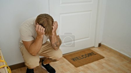 Distressed man crouching at home showing frustration, with a tattoo and welcome mat in the background.
