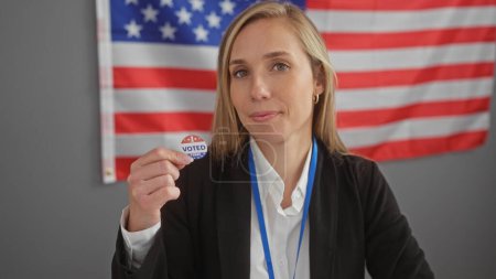 Confident young caucasian woman presents an 'i voted' sticker with american flag backdrop, symbolizing patriotic civic duty.