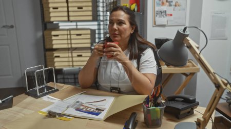 Photo for Middle-aged hispanic woman sips coffee in a cluttered detective's office, surrounded by paper, indoors. - Royalty Free Image
