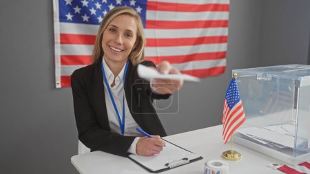 Una mujer caucásica sonriente extiende una boleta electoral en un colegio electoral americano con una bandera en el fondo.