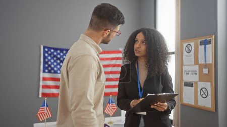 Un homme et une femme conversent dans un centre électoral américain avec des drapeaux américains, suggérant un cadre gouvernemental.