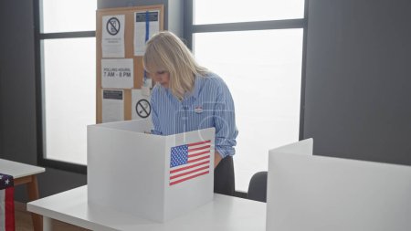 Téléchargez les photos : Une jeune femme blonde en chemise rayée dépose son bulletin de vote dans un bureau de vote américain, représentant la démocratie et le devoir civique. - en image libre de droit