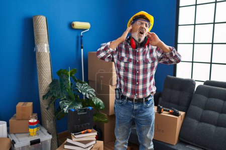Photo for Annoyed senior man covering ears with fingers from loud noise at new home, sporting hardhat and safety glasses - Royalty Free Image