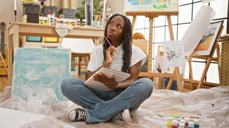 A contemplative woman artist sits cross-legged in a paint-splattered studio, pondering over a sketchpad.