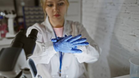 Foto de Una joven científica enfocada se pone guantes azules en un ambiente de laboratorio, insinuando investigación o trabajo médico. - Imagen libre de derechos