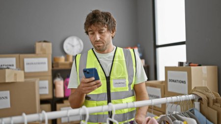 Photo for A man in a reflective vest labeled 'volunteer' uses a smartphone in a donation center storeroom. - Royalty Free Image