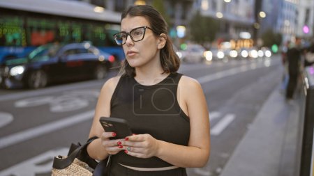 Foto de Hermosa mujer hispana morena en gafas, absorta en una conversación seria sobre teléfonos inteligentes en las calles urbanas de Madrid - Imagen libre de derechos