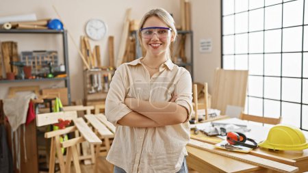 Confident young woman wearing safety glasses stands with arms crossed in a sunny carpentry workshop.
