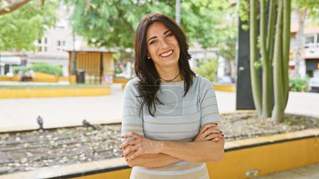 Mujer hispana madura sonriente con brazos cruzados de pie al aire libre en un parque verde de la ciudad.