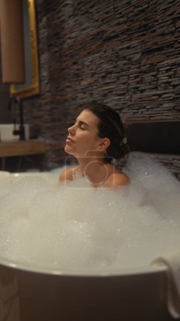 Foto de Mujer hispana joven relajándose sola en un lujoso baño interior de burbujas con los ojos cerrados, transmitiendo serenidad y autocuidado. - Imagen libre de derechos