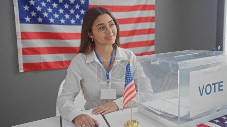 Foto de Trabajadora electoral hispana en centro de votación interior con bandera americana, sonriendo atractivamente. - Imagen libre de derechos