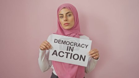 Selbstbewusste muslimische Frau mit einem "Demokratie in Aktion" -Schild vor rosa Hintergrund, das Aktivismus und Stärkung symbolisiert.