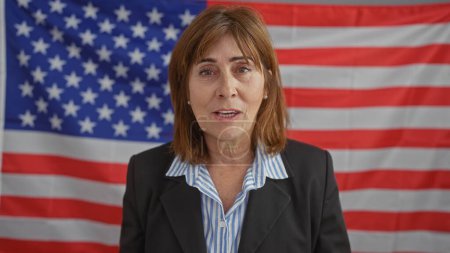 Foto de Mujer profesional de mediana edad con el pelo corto en un traje, posando con confianza delante de una bandera americana dentro de una oficina. - Imagen libre de derechos