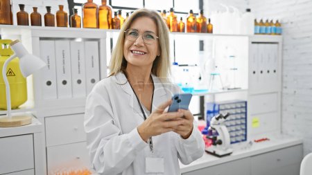 Une femme caucasienne souriante portant des lunettes et un manteau de laboratoire tenant un smartphone dans un cadre de laboratoire avec des étagères de bouteilles derrière elle.