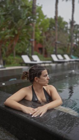 Foto de Una joven contemplativa se relaja en una lujosa piscina al aire libre en un complejo balinés, rodeado de follaje tropical. - Imagen libre de derechos