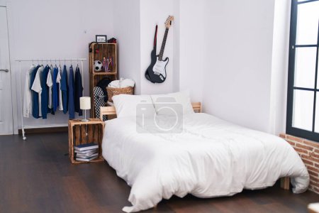 Photo for Cozy modern bedroom with natural light, a white bed, guitar on wall, and blue clothes on a rack. - Royalty Free Image