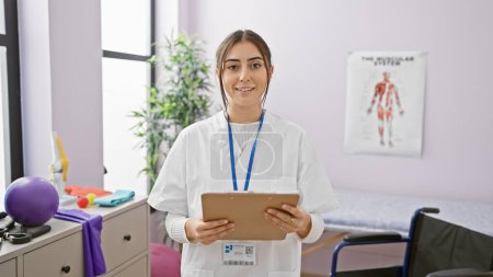 Téléchargez les photos : Jeune clinicienne hispanique tenant un presse-papiers se tient dans une salle de clinique de réadaptation, respirant le professionnalisme et la chaleur. - en image libre de droit