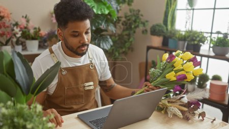 Photo for African american man working on laptop in flower shop surrounded by plants and floral arrangements - Royalty Free Image