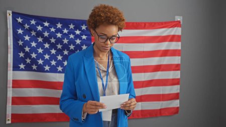 Foto de Mujer negra americana en chaqueta azul documento de lectura en el interior con nosotros bandera de fondo. - Imagen libre de derechos