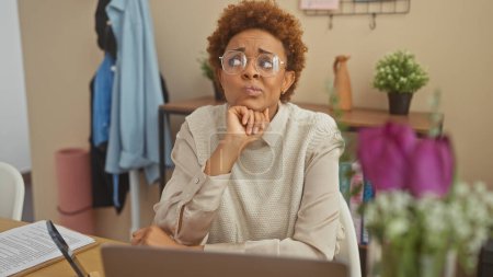 Pensive african american woman wearing glasses indoors beside her laptop in a cozy home setting.