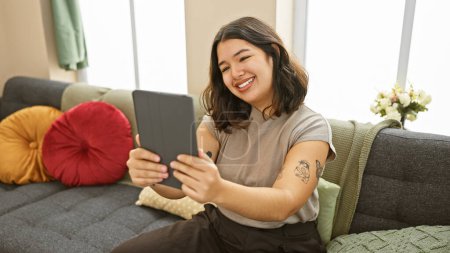 Téléchargez les photos : Jeune femme hispanique prenant un selfie avec une tablette à l'intérieur sur un canapé confortable entouré de coussins colorés. - en image libre de droit