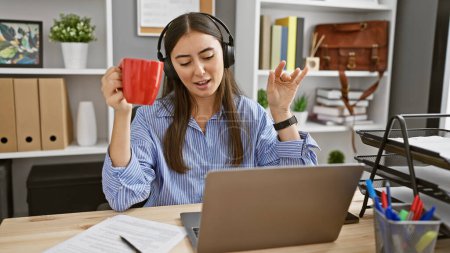 Foto de Una joven hispana disfruta del café mientras televisa, mostrando un moderno estilo de vida de trabajo remoto en una oficina en casa. - Imagen libre de derechos