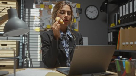 Téléchargez les photos : Femme blanche détective manger beignet au bureau du poste de police avec ordinateur portable, horloge et fichiers. - en image libre de droit