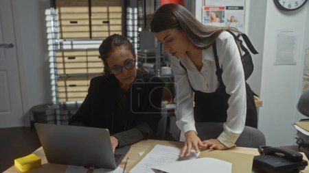 Téléchargez les photos : Deux femmes analysant des documents dans un bureau de poste de police, au milieu d'ordinateurs et de classeurs. - en image libre de droit