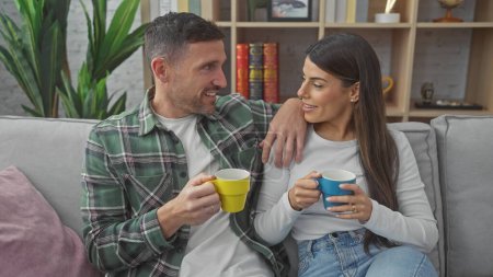 Foto de Un hombre y una mujer sonrientes sosteniendo cariñosamente tazas en un sofá en una acogedora sala de estar. - Imagen libre de derechos