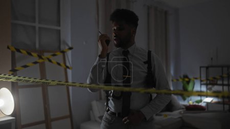 Foto de Un policía afroamericano en una habitación se comunica a través de walkie-talkie en una escena del crimen en el interior. - Imagen libre de derechos