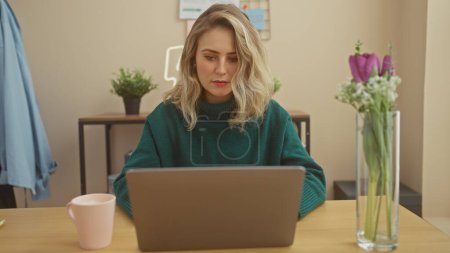 Téléchargez les photos : Une femme blonde concentrée travaille sur un ordinateur portable dans un cadre intérieur confortable, respirant un sentiment de professionnalisme décontracté. - en image libre de droit