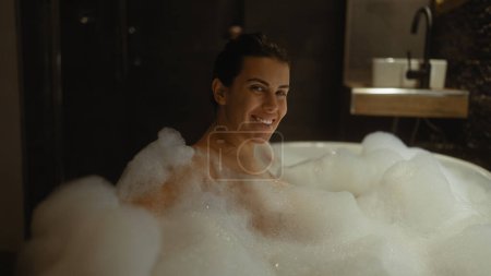 Foto de Una joven sonriente disfruta de un relajante baño de burbujas en un entorno de baño interior. - Imagen libre de derechos