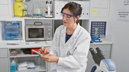 Photo for Hispanic woman scientist using smartphone in laboratory setting, likely reviewing data or communicating. - Royalty Free Image