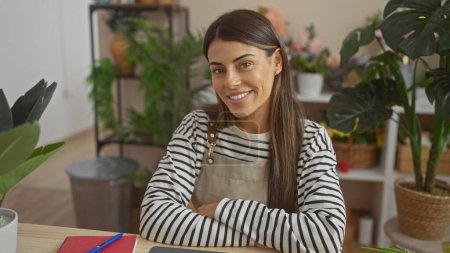 Téléchargez les photos : Smiling hispanic woman with crossed arms wearing an apron in a plant-filled living room. - en image libre de droit